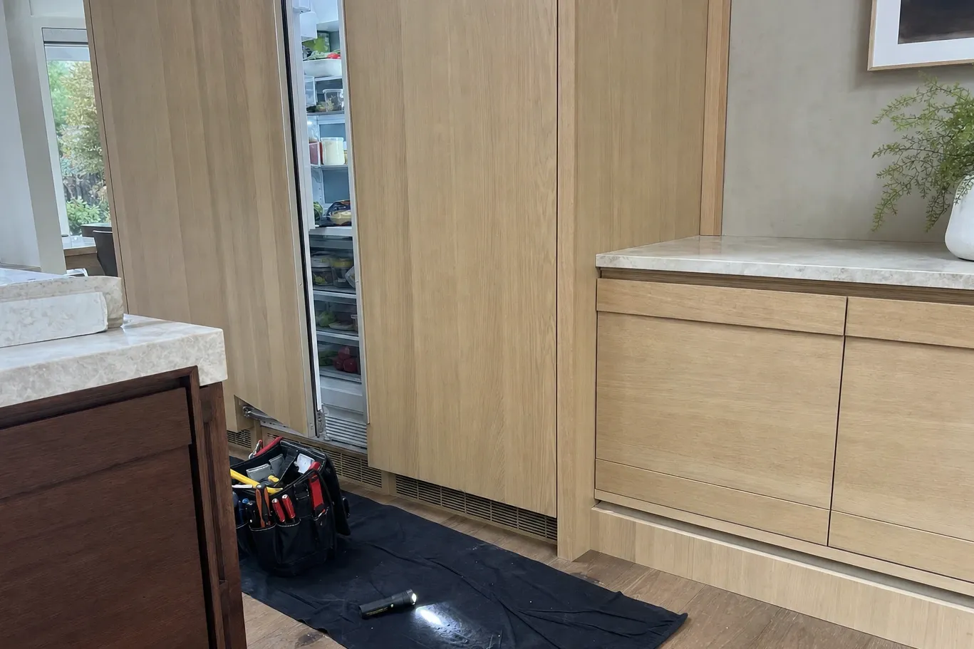 Panel-ready refrigerator column behind oak cabinet fronts, door ajar on stocked shelves, tool bag and flashlight on a drop cloth